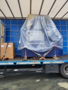 Large machinery securely covered and strapped on a truck for transport, alongside cardboard boxes.